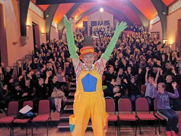 Caroline Ainslie dressed in yellow dungarees and green gloves in front of an audience of children